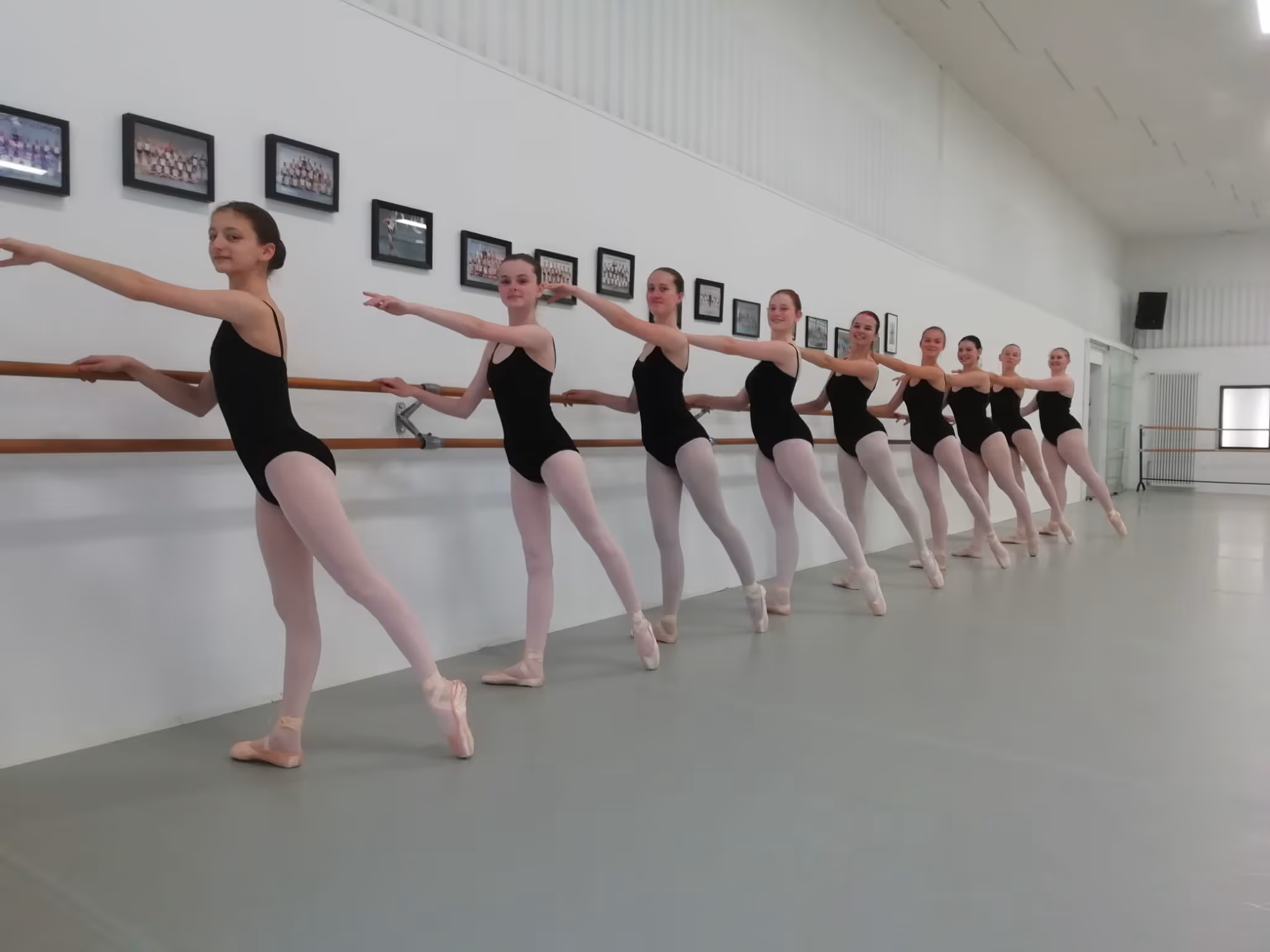 Eine Gruppe von neun Balletttänzerinnen in schwarzem Trikot und rosa Strumpfhosen trainiert an einer Ballettstange in einem hellen Tanzstudio.