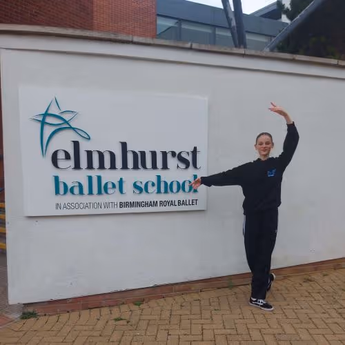 Tänzerin posiert in schwarzer Kleidung neben einem Schild der Elmhurst Ballet School an einer weißen Wand.