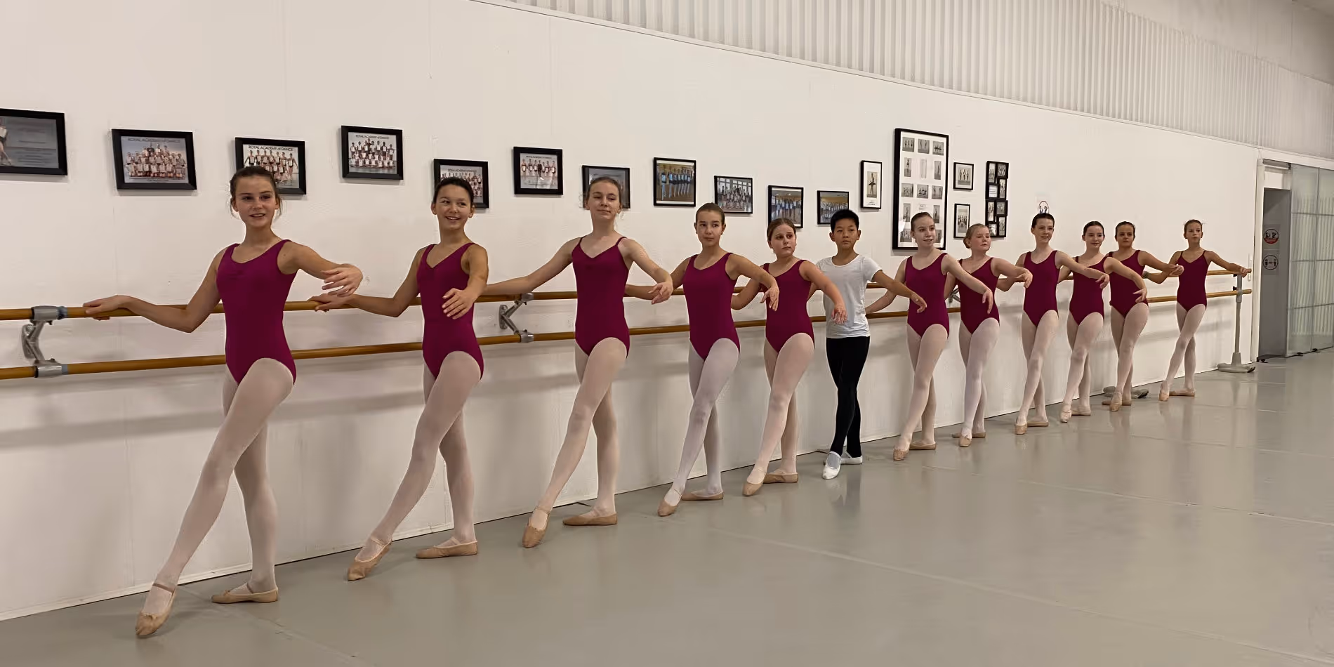 Zwölf Ballettschüler stehen in einer Reihe an einer Ballettstange in einem Tanzstudio, elf Mädchen in magentafarbenen Trikots und ein Junge in einem weißen Shirt und schwarzer Hose.