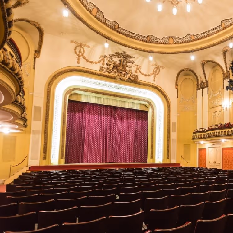 Innenraum eines Theaters mit roten Sitzreihen, einer Bühne mit rotem Vorhang und kunstvoll verzierten Wänden und Decke.